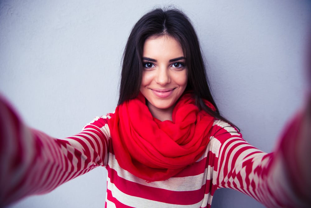 Happy cute woman making selfie over gray background. Wearing in bright scarf and sweater. Looking at camera